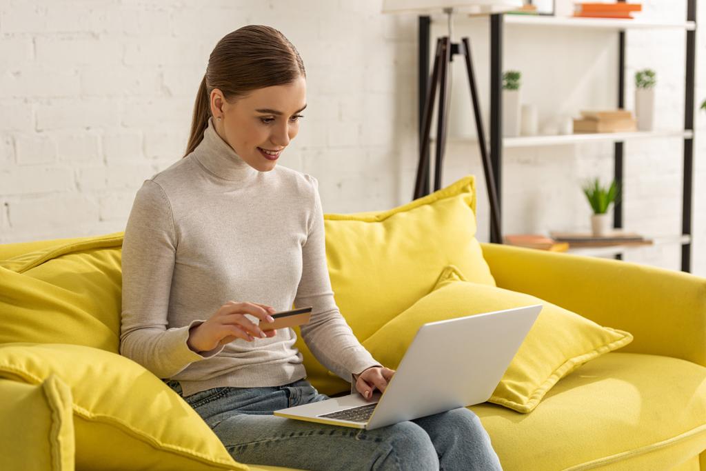 Houkutteleva hymyilevä nainen, jolla on luottokortti ja käyttää kannettavaa tietokonetta sohvalla
 - Valokuva, kuva