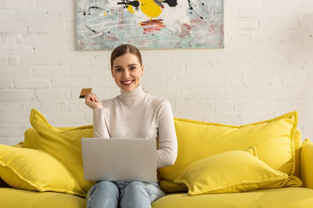 Hermosa chica sonriendo a la cámara y sosteniendo la tarjeta de crédito con el ordenador portátil en el sofá
 - Foto, Imagen