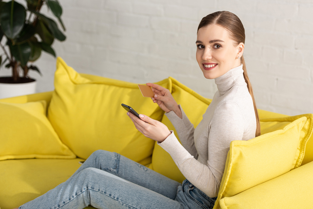 Gyönyörű nő mosolyog a kamera használata közben okostelefon és a kezében hitelkártya kanapén - Fotó, kép