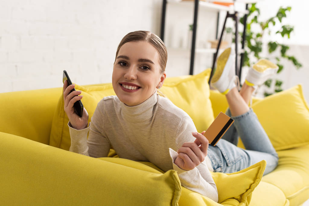 Mujer sonriente sosteniendo teléfono inteligente y tarjeta de crédito mientras está acostada en el sofá
 - Foto, Imagen