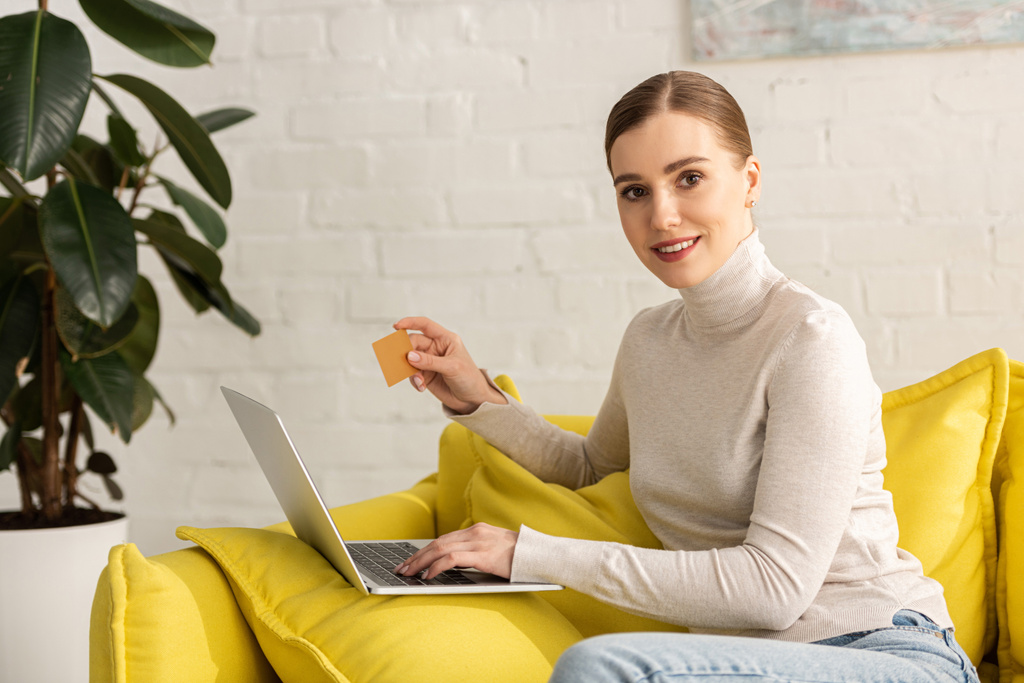 Atrakcyjna kobieta uśmiechnięta do kamery podczas korzystania z laptopa i karty kredytowej na kanapie w salonie - Zdjęcie, obraz
