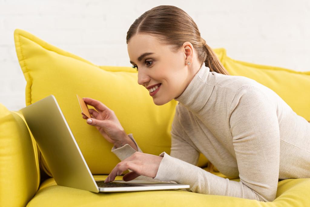 Vista lateral de la mujer sonriente usando el ordenador portátil y la celebración de la tarjeta de crédito mientras está acostado en el sofá
  - Foto, imagen