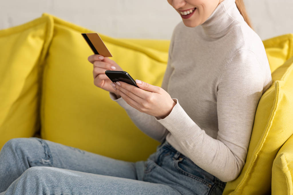 Przycięty widok uśmiechniętej dziewczyny za pomocą smartfona i trzymając kartę kredytową na kanapie - Zdjęcie, obraz
