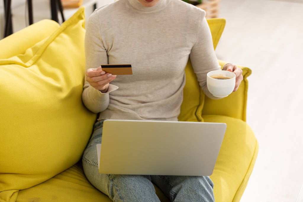 Cropped view of woman with credit card and laptop drinking coffee on couch - Photo, Image