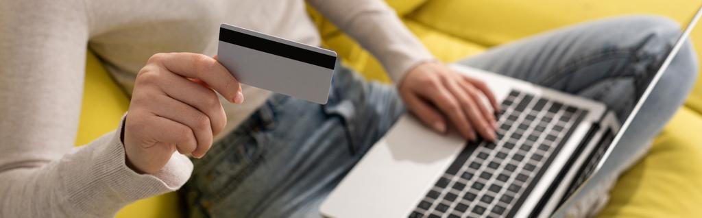 Widok na dziewczynę pokazującą kartę kredytową i korzystającą z laptopa na sofie, panoramiczne ujęcie - Zdjęcie, obraz
