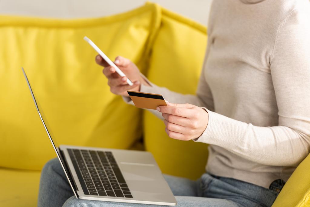 Обрізаний вид дівчини, що тримає кредитну картку, смартфон і ноутбук на дивані
 - Фото, зображення