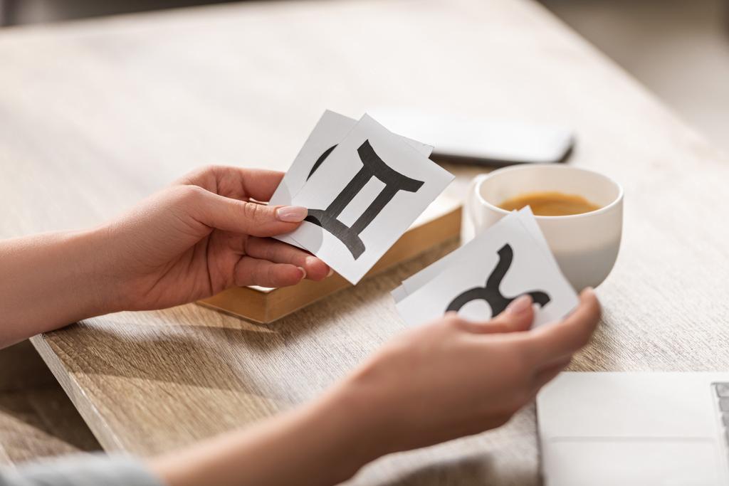 Oříznutý pohled na astrolog držící karty se zvěrokruhovými znaky vedle kávy a knihy na stole - Fotografie, Obrázek