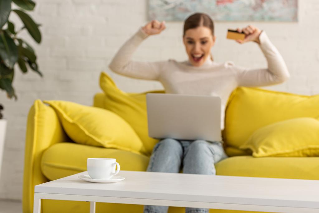 Válogatott fókusz kávé asztalra és izgatott nő hitelkártyával és laptop kanapén a háttérben - Fotó, kép