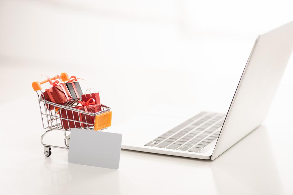Toy cart with gift boxes beside credit card and laptop on white surface - Photo, Image