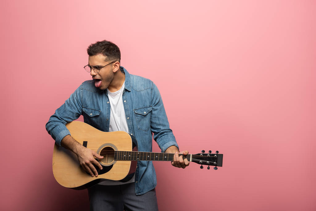 Mladý muž vyčnívat jazyk při hraní na akustické kytary na růžovém pozadí - Fotografie, Obrázek