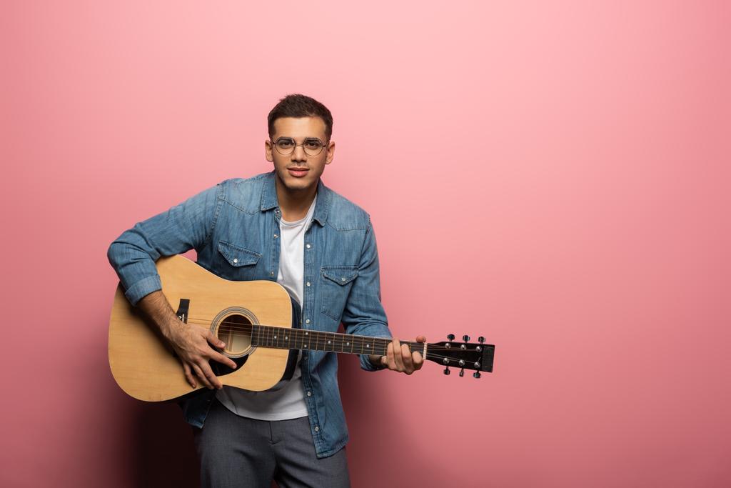 Knappe man op zoek naar camera tijdens het spelen van gitaar op roze achtergrond - Foto, afbeelding