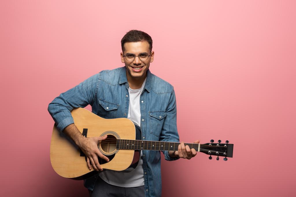 Young man smiling at camera and playing acoustic guitar on pink background - Photo, Image