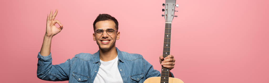 Panoramic shot of man with acoustic guitar smiling at camera and showing okay isolated on pink  - Photo, Image