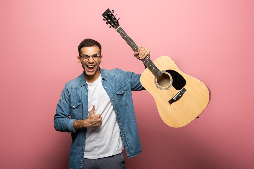 Cheerful man with acoustic guitar showing thumb up sign on pink background - Photo, Image