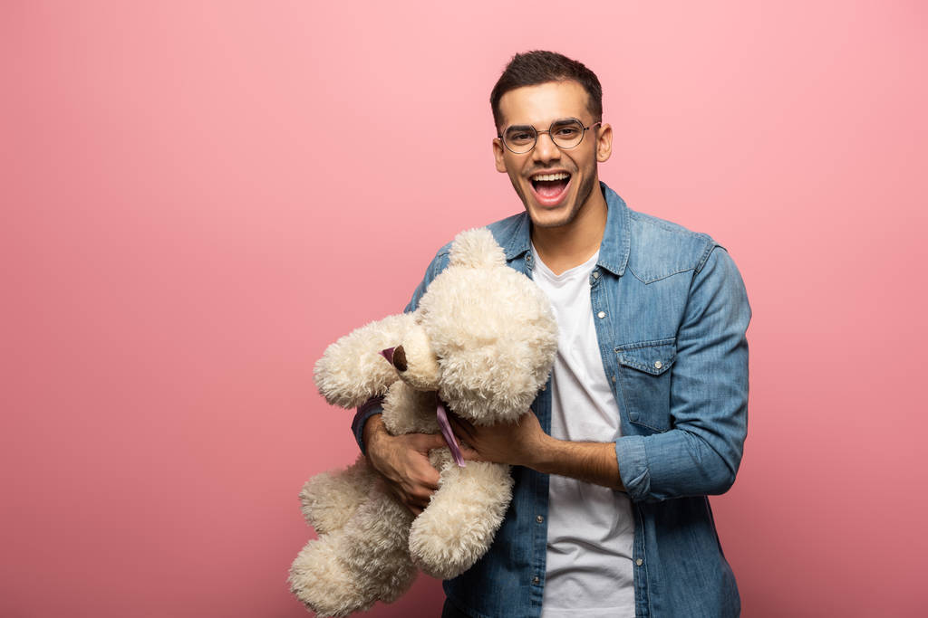 Vrolijke jonge man houden teddy beer en kijken naar de camera op roze achtergrond - Foto, afbeelding