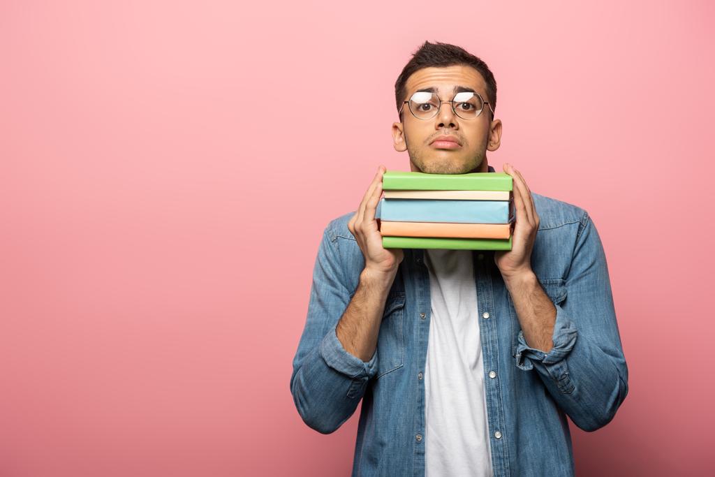 Запутавшийся молодой человек держит красочные книги и смотрит в камеру на розовом фоне
 - Фото, изображение
