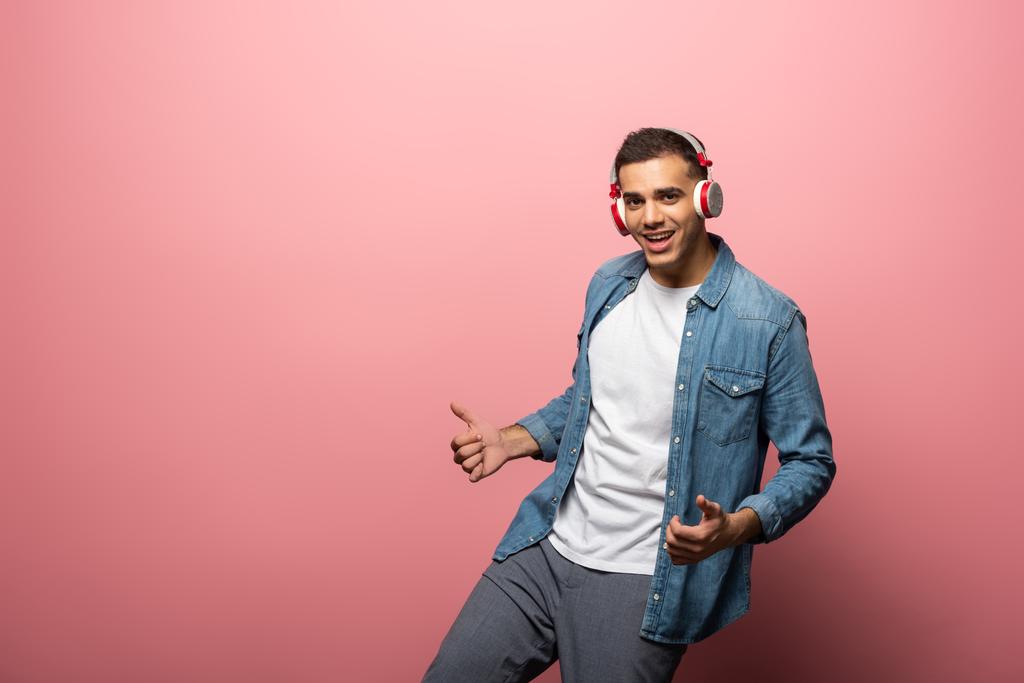 Fiatal mosolygós férfi fülhallgató mutatja hüvelykujj fel jel rózsaszín háttér - Fotó, kép