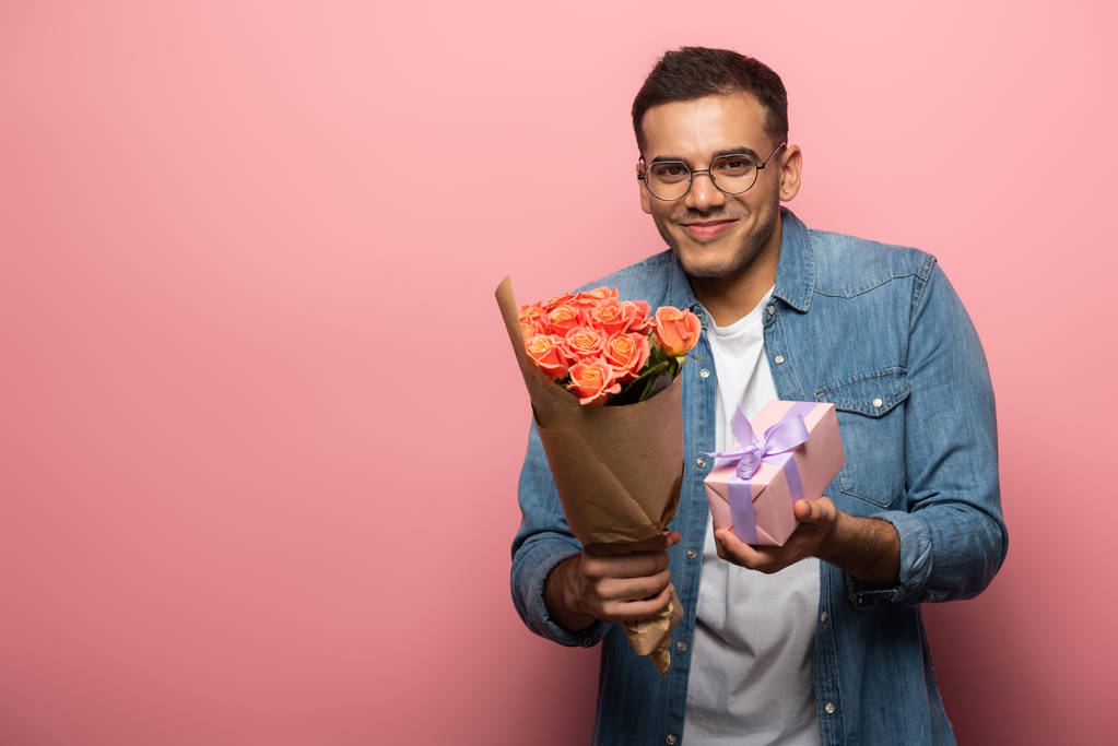 Young man smiling at camera while holding present and bouquet on pink background - 写真・画像
