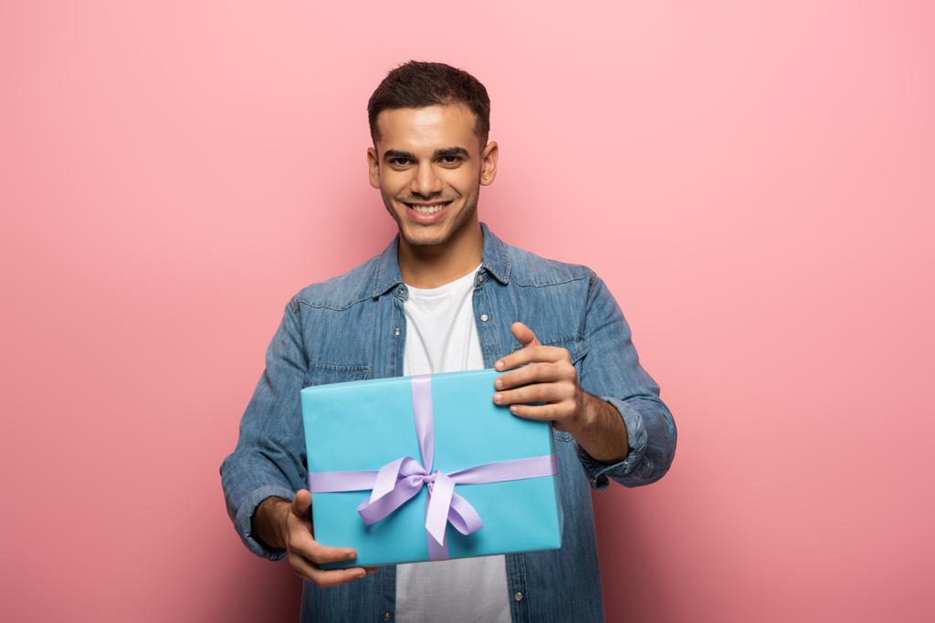Молодий чоловік посміхається на камеру, тримаючи подарунок на рожевому фоні
 - Фото, зображення