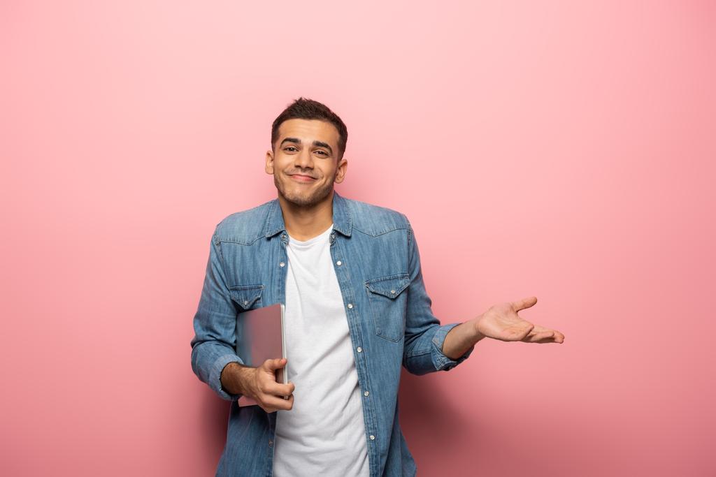 Junger lächelnder Mann mit Laptop zeigt Achselzucken-Geste auf rosa Hintergrund - Foto, Bild