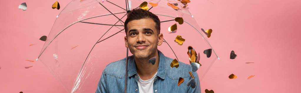 Pembe konfeti altında şeffaf bir şemsiyeyle gülümseyen adamın panoramik görüntüsü. - Fotoğraf, Görsel