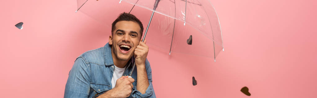 Panoramic shot of cheerful man with transparent umbrella under falling confetti on pink background  - Photo, Image