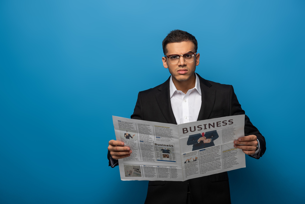 Pensive businessman looking at camera while holding newspaper on blue background - Photo, Image