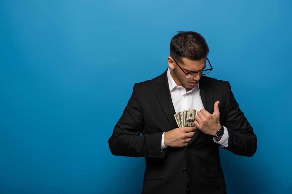 Бизнесмен кладет долларовые купюры в карман пиджака на синем фоне
 - Фото, изображение
