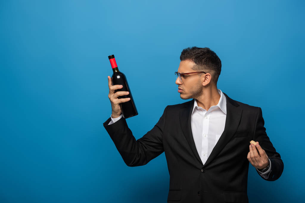 Vista lateral do empresário segurando garrafa de vinho e cortiça no fundo azul
 - Foto, Imagem