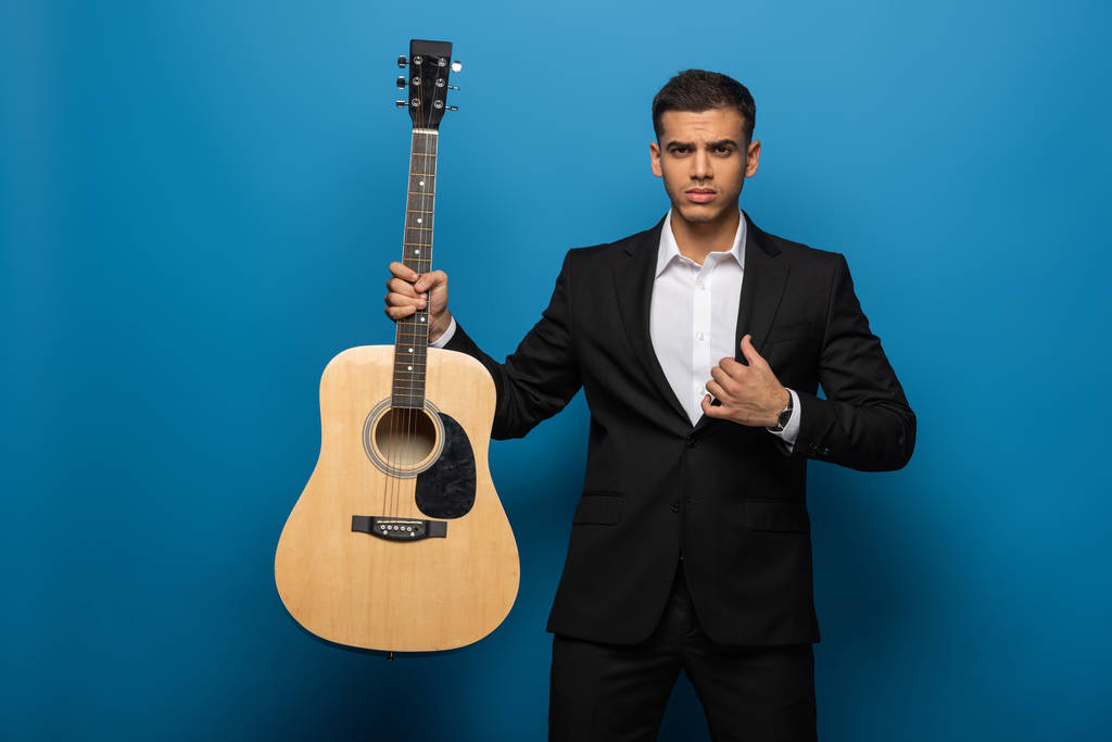 Красивый бизнесмен держит акустическую гитару и смотрит на камеру на синем фоне
 - Фото, изображение