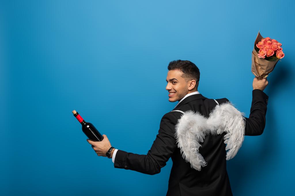 Back view of smiling businessman with white wings holding wine bottle and bouquet on blue background - Photo, Image
