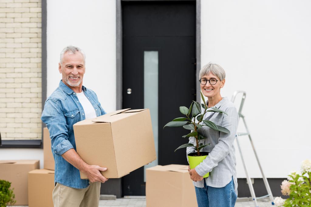 Älterer Mann hält Schachtel und lächelnde Frau hält Pflanze in der Nähe des neuen Hauses  - Foto, Bild