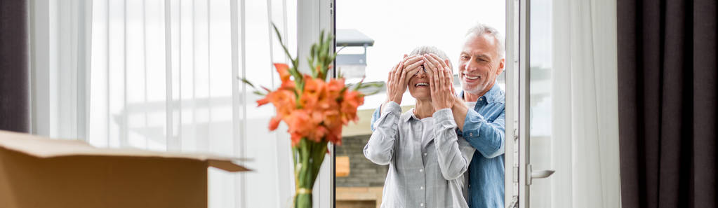 panoramisch schot van glimlachende man verduisteren gezicht van vrouw in nieuw huis  - Foto, afbeelding