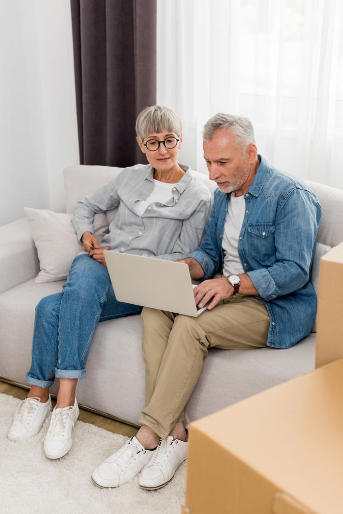 Dojrzały mężczyzna za pomocą laptopa i kobieta patrząc na to w nowym domu  - Zdjęcie, obraz