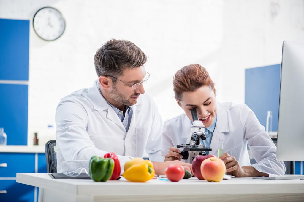 nutricionista molecular sonriente usando microscopio y su colega mirándola
 - Foto, Imagen