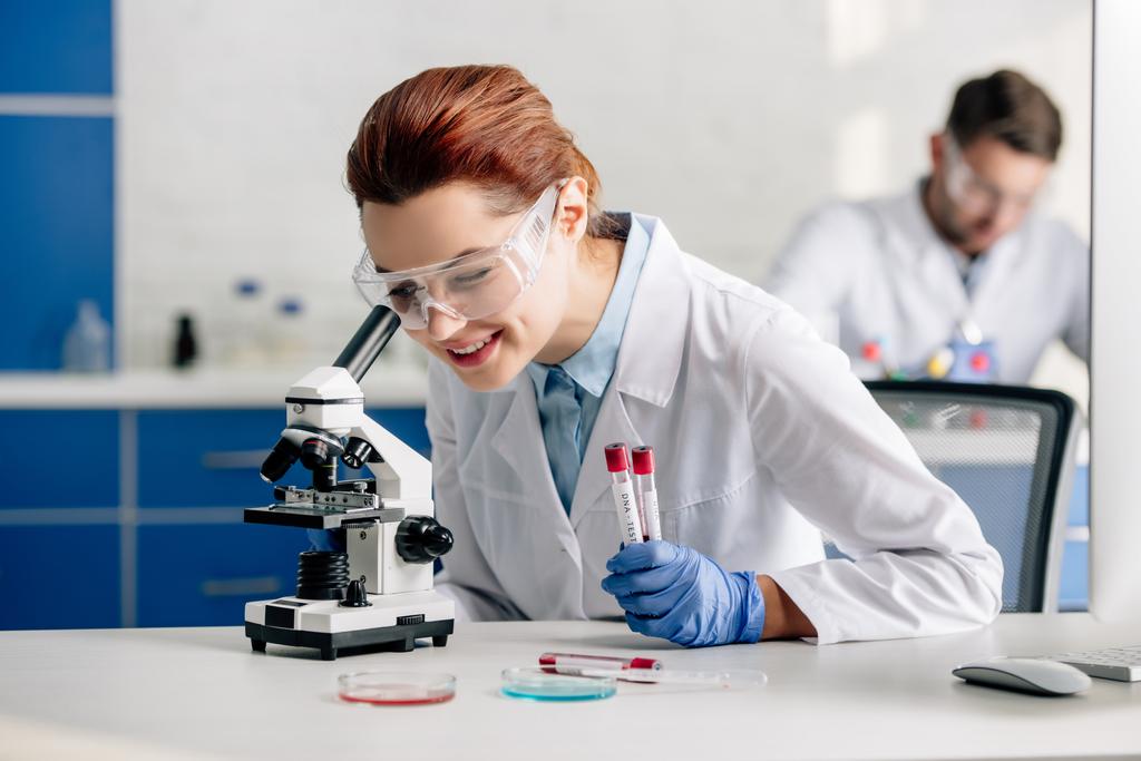 selektywne skupienie się uśmiechniętego konsultanta genetycznego wykonującego badania DNA i stosującego mikroskop w laboratorium  - Zdjęcie, obraz