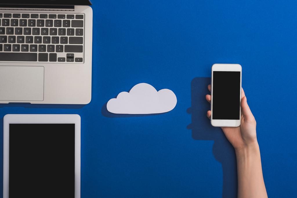 cropped view of woman holding smartphone near empty white paper cloud and laptop with digital tablet on blue - Photo, Image