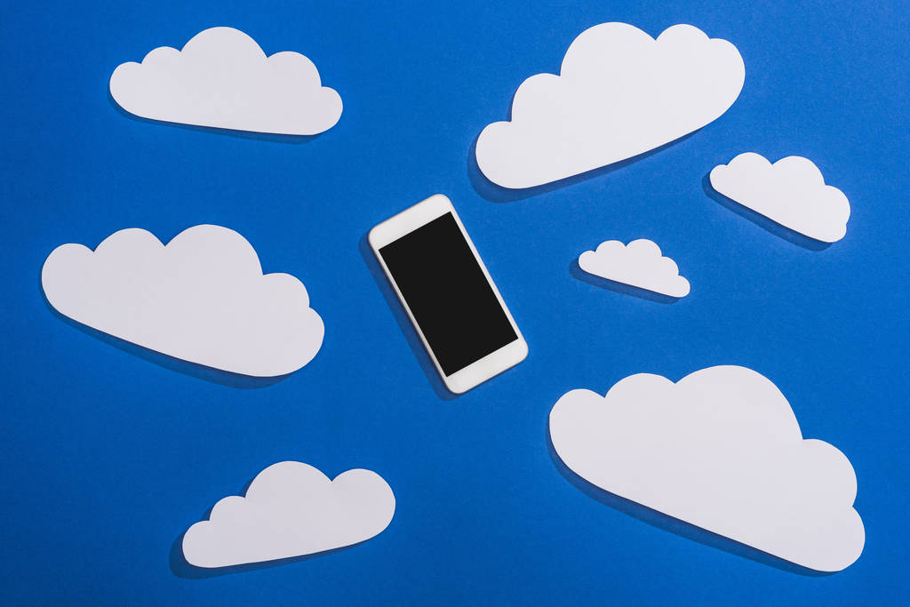 πάνω όψη του λευκού χαρτιού κομμένα σύννεφα και smartphone σε μπλε φόντο - Φωτογραφία, εικόνα