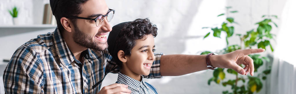 панорамный снимок улыбающегося еврейского отца, указывающего пальцем и смотрящего в окно с сыном в квартире
  - Фото, изображение