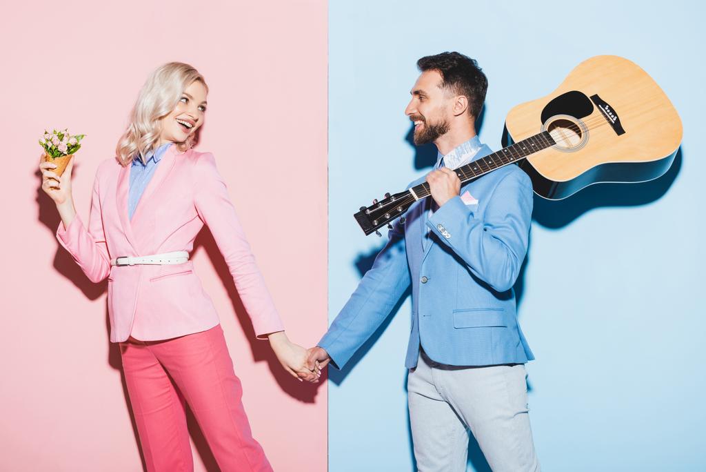 glimlachende vrouw met boeket en knappe man met akoestische gitaar hand in hand op roze en blauwe achtergrond  - Foto, afbeelding