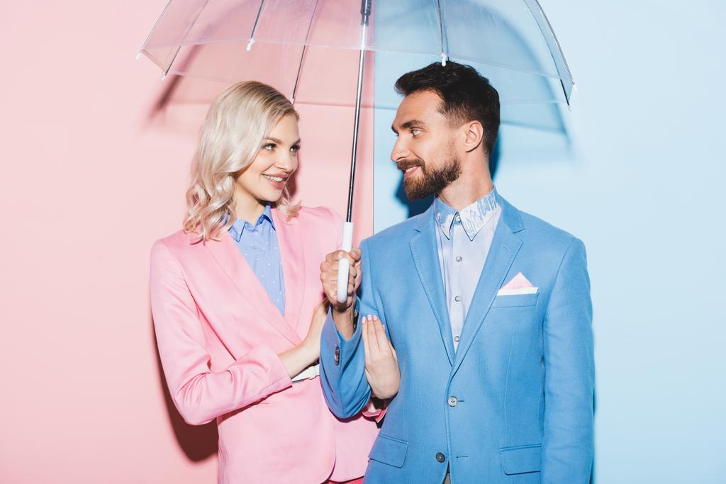 glimlachende vrouw en knappe man met paraplu op roze en blauwe achtergrond  - Foto, afbeelding