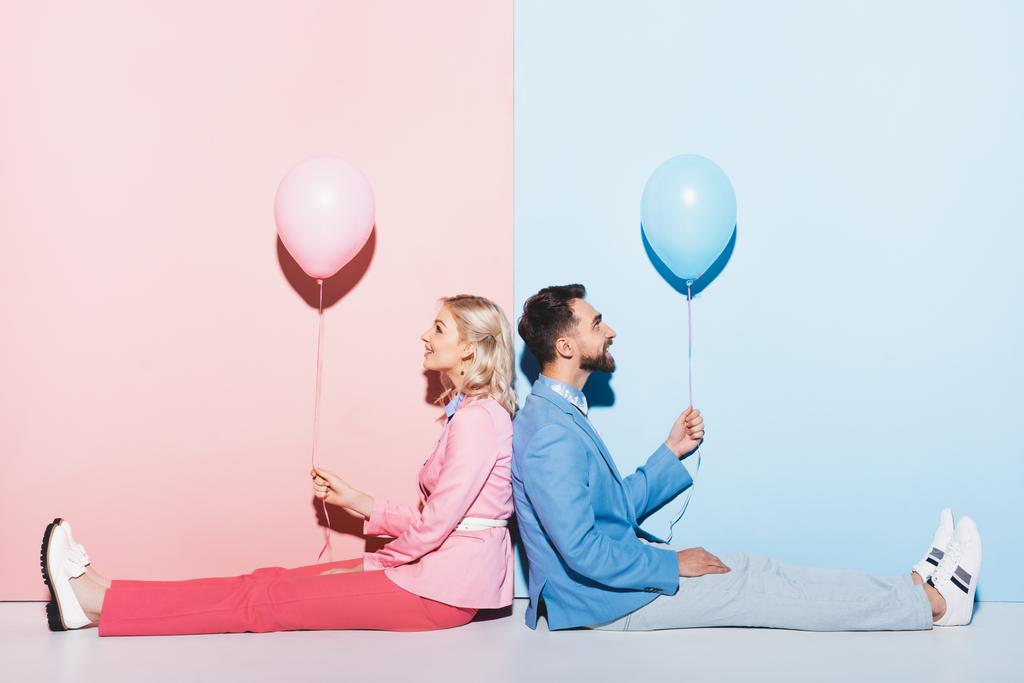 back to back view of smiling woman and handsome man holding balloons on pink and blue background  - Photo, Image