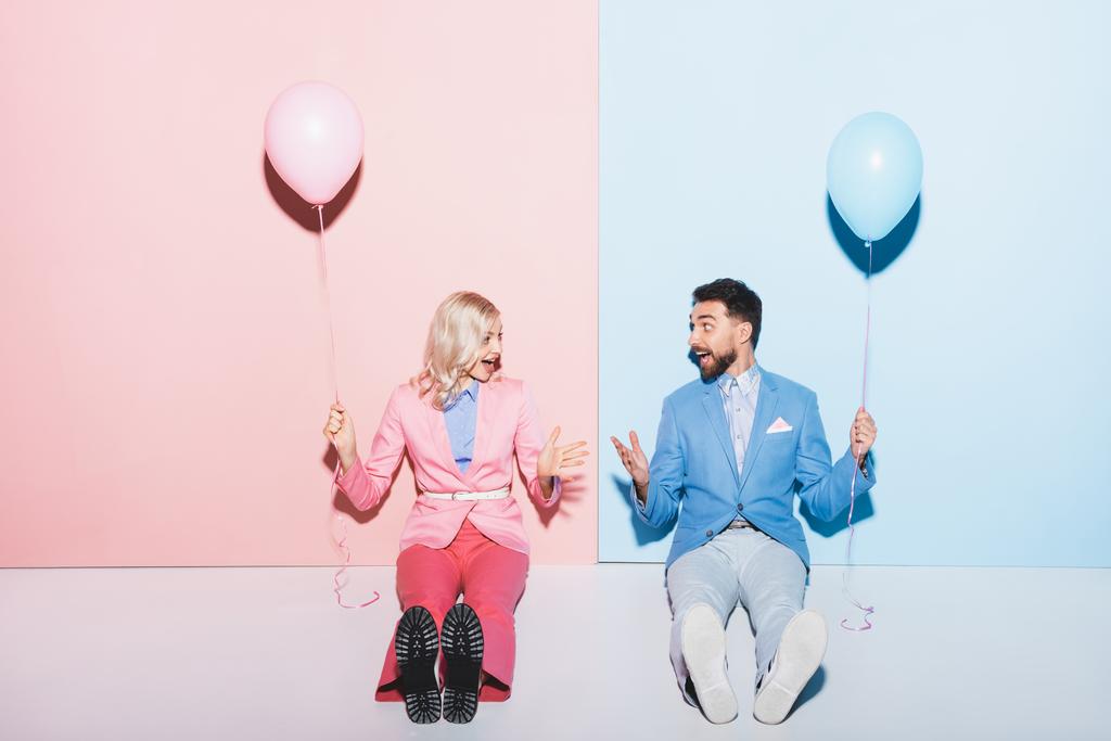 σοκαρισμένη γυναίκα και όμορφος άντρας κρατώντας μπαλόνια σε ροζ και μπλε φόντο  - Φωτογραφία, εικόνα