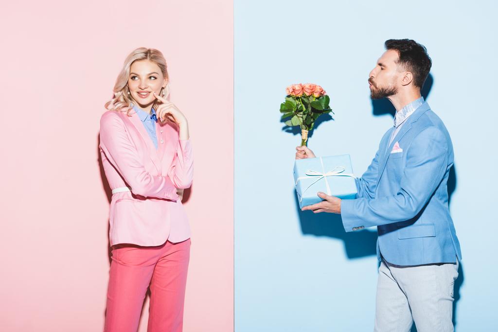 όμορφος άντρας δίνει σε ονειρεμένη γυναίκα μπουκέτο και δώρο σε ροζ και μπλε φόντο  - Φωτογραφία, εικόνα