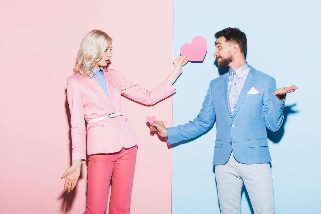 geschokte vrouw en knappe man met hartvormige kaarten op roze en blauwe achtergrond  - Foto, afbeelding