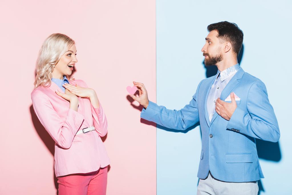 knappe man geeft hartvormige kaart aan geschokte vrouw op roze en blauwe achtergrond  - Foto, afbeelding
