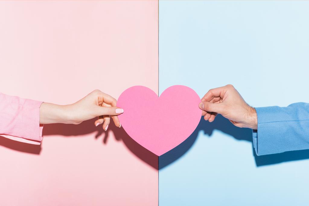 bijgesneden weergave van man en vrouw met hartvormige kaart op roze en blauwe achtergrond  - Foto, afbeelding