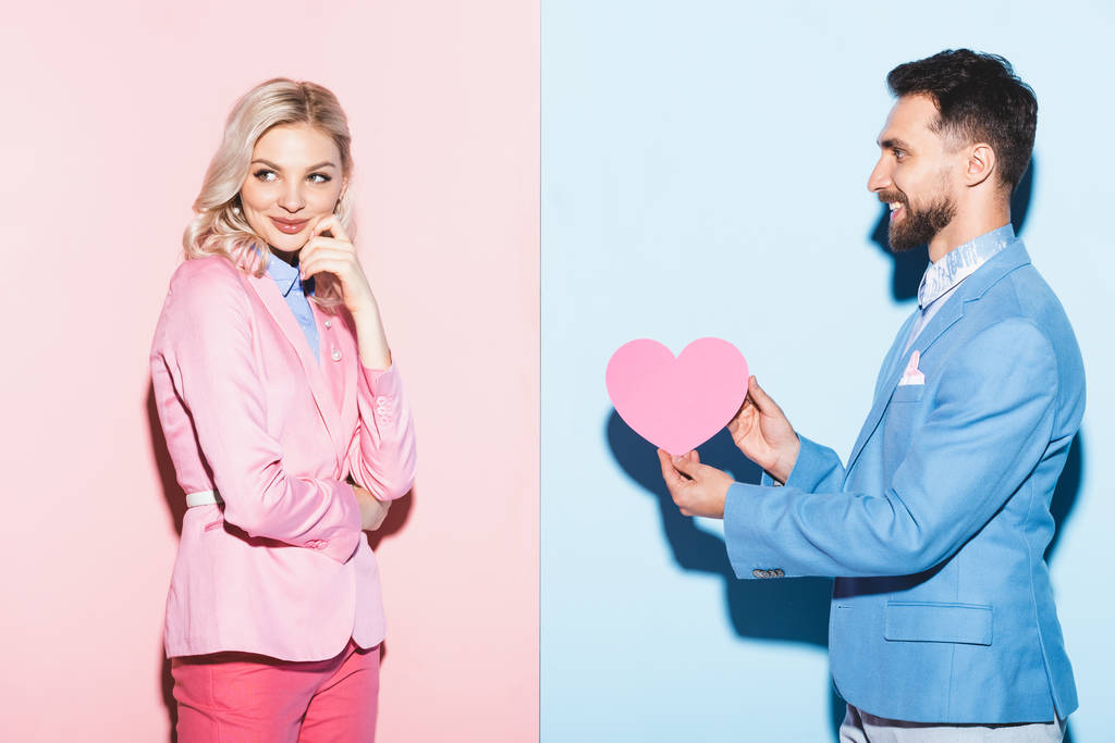 Красивый мужчина дарит открытку в форме сердца мечтательной женщине на розовом и синем фоне
  - Фото, изображение