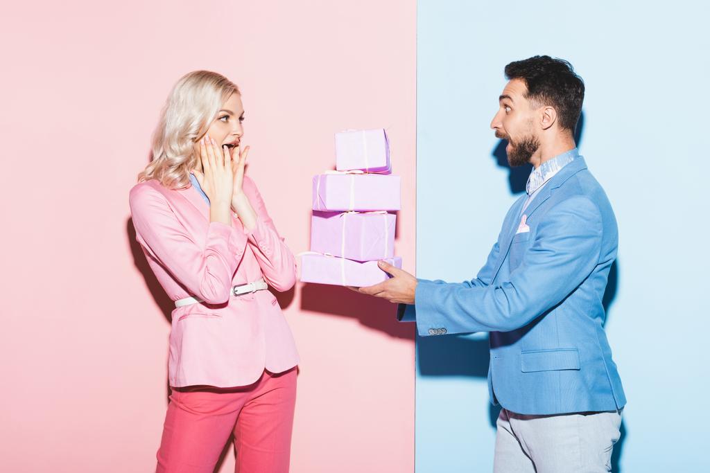 όμορφος άνδρας δίνει δώρα σε σοκαρισμένη γυναίκα σε ροζ και μπλε φόντο  - Φωτογραφία, εικόνα