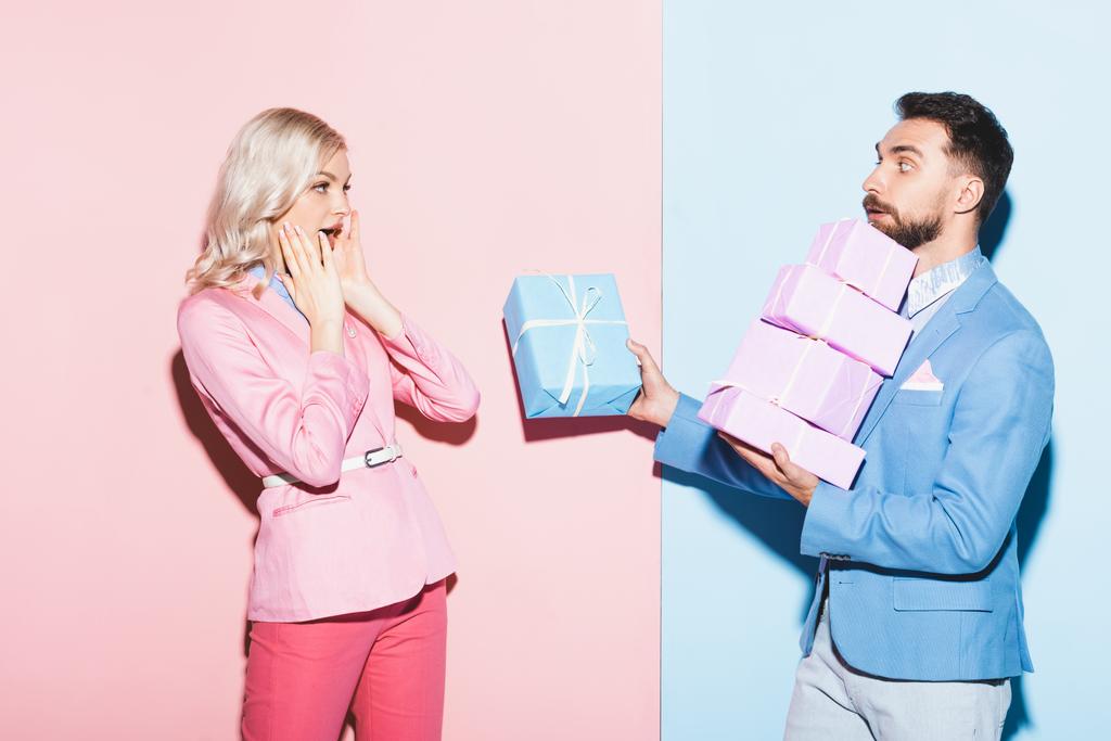pohledný muž dává dárek šokované ženy na růžovém a modrém pozadí  - Fotografie, Obrázek